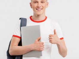 Blonder junger Mann in weißem T-Shirt mit Laptop und Rucksack zeigt "Daumen hoch!"