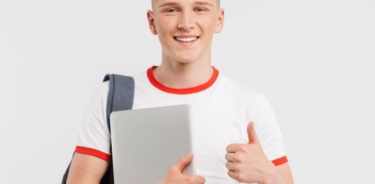 Blonder junger Mann in weißem T-Shirt mit Laptop und Rucksack zeigt "Daumen hoch!"