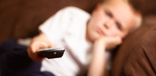 Ein Junge auf dem Sofa liegend richtet eine ernbedienunge aus dem Bild heraus.