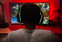 EIn junge in weißem T-Shirt mit dicken Kopfhörerohrmuscheln von hinten s´gesehen schaut auf einen Bildschirm vor einer rot erleuchteten Wnad.