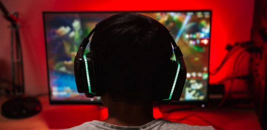 EIn junge in weißem T-Shirt mit dicken Kopfhörerohrmuscheln von hinten s´gesehen schaut auf einen Bildschirm vor einer rot erleuchteten Wnad.