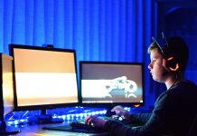 Ein Junge mit dicken Kopfhörerrn vor drei Computerbildschirmen im Dunkeln