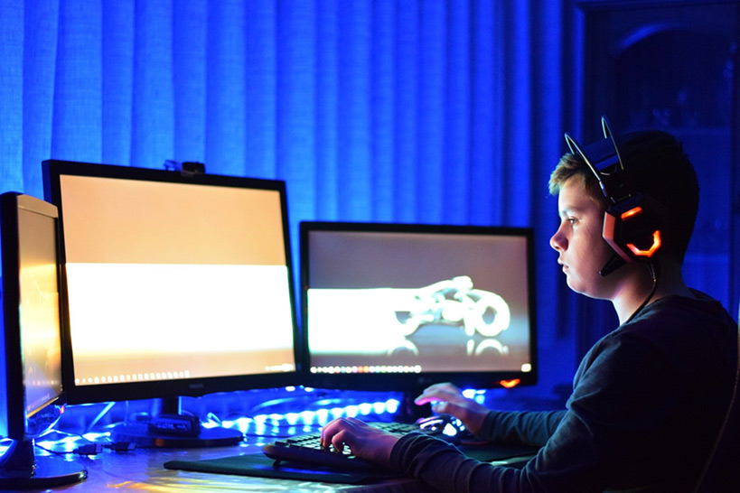 Ein Junge mit dicken Kopfhörerrn vor drei Computerbildschirmen im Dunkeln