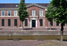 Schullandschaft im Wandel: Bürgerentscheid in Ostfriesland sorgt für Unsicherheit Ein relativ niedriges, breites zweistöckiges Backsteingebäude aus dem 19. Jahrhundert mit hervorgehobenem Portikus an einer klinkergepflasterten Straße hinter einem kleinen Kanal, davor drei Bäume.