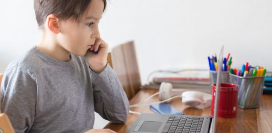 Ein junge im grauen Pullover sitzt inmitten von SChulutensilien am Laptop.