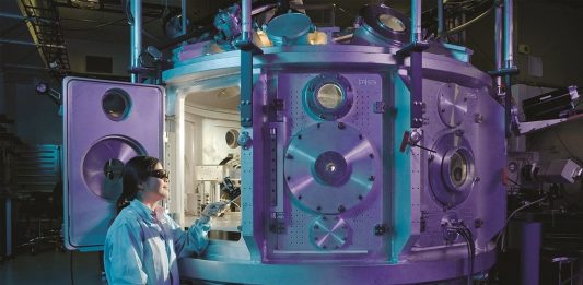 Eine Frau mit Schutzbrille gekleidet in einen weißen Kittel schaut in die geöffnetet Tür einer technischen ApparaturKittel