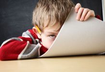 Fernunterricht läuft weiter – und unter Eltern reift die Erkenntnis, dass das Unterrichten potenziell unwilliger Schüler ein ganz schön harter Job ist Ein Junge, vor einer Schultafel auf seinen Tisch gesunken, schaut an seinem halb heraufgezogenen Schreibblock vorbei.