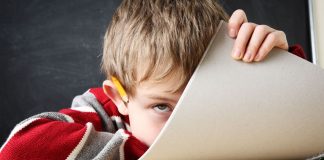 Ein Junge, vor einer Schultafel auf seinen Tisch gesunken, schaut an seinem halb heraufgezogenen Schreibblock vorbei.