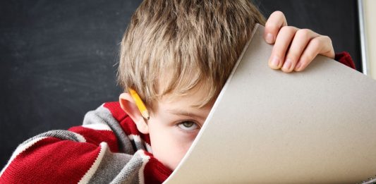 Ein Junge, vor einer Schultafel auf seinen Tisch gesunken, schaut an seinem halb heraufgezogenen Schreibblock vorbei.