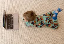 Kleinkind im SChlafanzug auf einem Sisalteppich bäuchlings vor einem Laptop