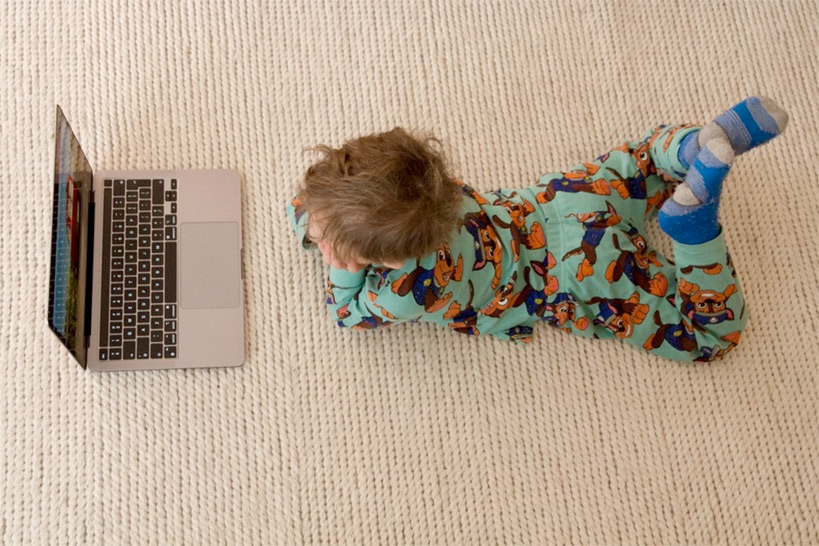 Kleinkind im SChlafanzug auf einem Sisalteppich bäuchlings vor einem Laptop