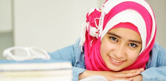 junges Mädchen mit rot-weiß gestreiftem Kopftuch über Bücher gebeugt blickt lächelnd auf