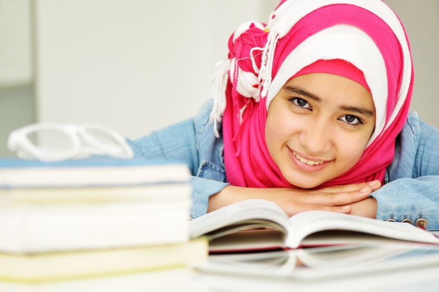 junges Mädchen mit rot-weiß gestreiftem Kopftuch über Bücher gebeugt blickt lächelnd auf
