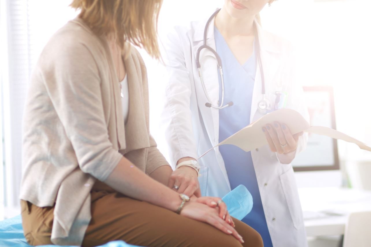 Eine PAtientin sitzt quer auf einer Behandlungsliege, von der Seite fasst eine Ärztin an ihren Puls während sie die auf einem Diagnosezettel geschriebene Behandlung erläutert.
