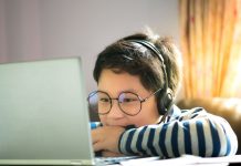 junge mit Brille und Kopfhörern in gestreiftem Pullover blickt über eine TIschplatte hinweg auf ein Laptop