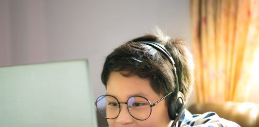 junge mit Brille und Kopfhörern in gestreiftem Pullover blickt über eine TIschplatte hinweg auf ein Laptop