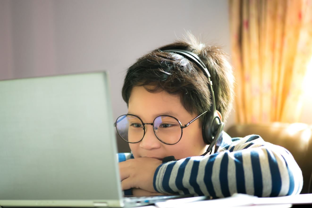 junge mit Brille und Kopfhörern in gestreiftem Pullover blickt über eine TIschplatte hinweg auf ein Laptop