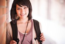 Eine junge Frau mit schulterlangem dunklem Haar und Ohrringen, die einen Rucksack trägt und lächelnd in die Kamera schaut.