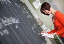 Eine Mathelehrerin schreibt, von einem Blatt ablesend Formeln auf eine Tafel.