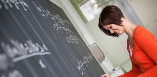 Eine Mathelehrerin schreibt, von einem Blatt ablesend Formeln auf eine Tafel.