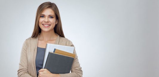 lächelnde Junge Frau mit Heften und eienn Block im Arm vor grauem Hintergrund