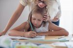 Frontalansicht einer blonde Frau beugt sich von hinten über ein amm Tisch sitzendes Mädchen mit Stift und Heft - Homeschooling stellt auch Eltern vor neue Aufgaben. Foto: Shutterstock