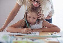 Homeschooling-Studie: Gelungener Heimunterricht aus Sicht der Eltern Frontalansicht einer blonde Frau beugt sich von hinten über ein amm Tisch sitzendes Mädchen mit Stift und Heft - Homeschooling stellt auch Eltern vor neue Aufgaben. Foto: Shutterstock