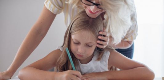 Frontalansicht einer blonde Frau beugt sich von hinten über ein amm Tisch sitzendes Mädchen mit Stift und Heft - Homeschooling stellt auch Eltern vor neue Aufgaben. Foto: Shutterstock