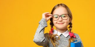 Ein lächelndes Mädchen mit zwei Zöpfen und Brille steht vor einem gelben Hintergrund. Sie trägt einen grauen Pullover und hält ein blaues Buch in der Hand.