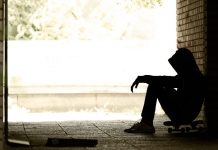 Jugendlicher in Hoodie, sitzt im Schatten eng zusammengekauert an eine Mauer gelehnt.