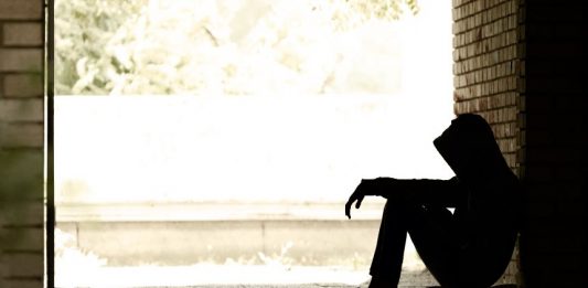 Jugendlicher in Hoodie, sitzt im Schatten eng zusammengekauert an eine Mauer gelehnt.