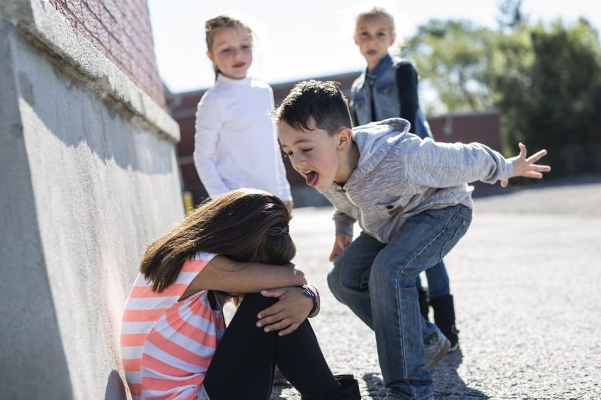 Ein Junge schreit ein an einer Wand hockendes Mädchen an. Im Hintergrund schauen zwei Kinder zu.