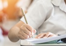 Landesschülerrat: Bedingungen für Wechselunterricht Schule überlassen Großaufnahme der Hände einer Person in einem weißen Poloshirt in einer Prüfung, die ein Blatt auf ein Pult drückt und mit einem Stift darauf schreibt. Über die rechte Schulter ist ein unscharfer Hintergrund mit weiteren weißgekleideten Personen an Tischen erkennbar, die in einer Turnhalle platziert zu sein scheinen.