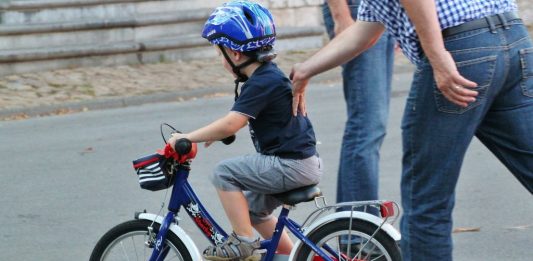 EIn Mann in kariertem Hemd schiebt ein kleines Kind auf dem Fahrrad an.