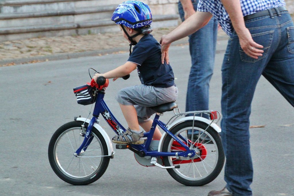 EIn Mann in kariertem Hemd schiebt ein kleines Kind auf dem Fahrrad an.