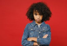 Ein Mädchen mit dunklen, krausen Haaren steht mit verschränkten Armen vor einem roten Hintergrund.