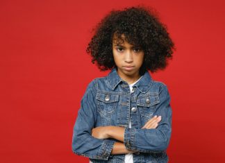 Studie zeigt verbreiteten Rassismus – Neues Gesetz soll besser vor Diskriminierung schützen (auch durch Schulen) Ein Mädchen mit dunklen, krausen Haaren steht mit verschränkten Armen vor einem roten Hintergrund.