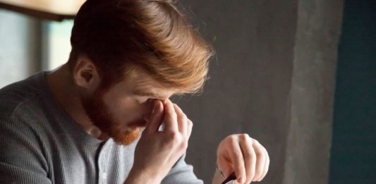 Junger Mann mit Brille in der Hand am Schreibtisch massiert seine Nasenwurzel.