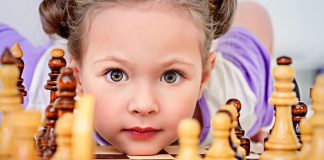 Ein Kindergartenkind schaut hinter einem Schachbrett liegend durch Schachfiguren hindurch.