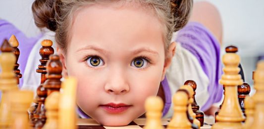 Ein Kindergartenkind schaut hinter einem Schachbrett liegend durch Schachfiguren hindurch.