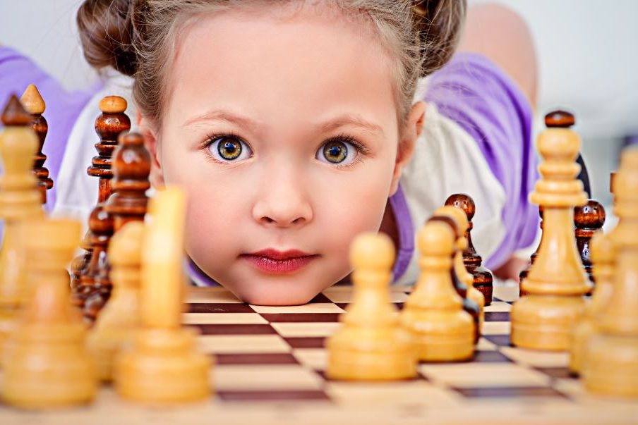 Ein Kindergartenkind schaut hinter einem Schachbrett liegend durch Schachfiguren hindurch.