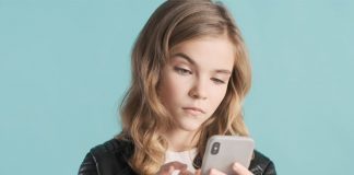 Soziale Medien: Aufwärtsvergleiche mindern das Wohlbefinden von Kindern und Jugendlichen Langhaariges Mädchen vor blauem Hintergrund tippt mit skeptischem Blick auf ihr Smartphone.