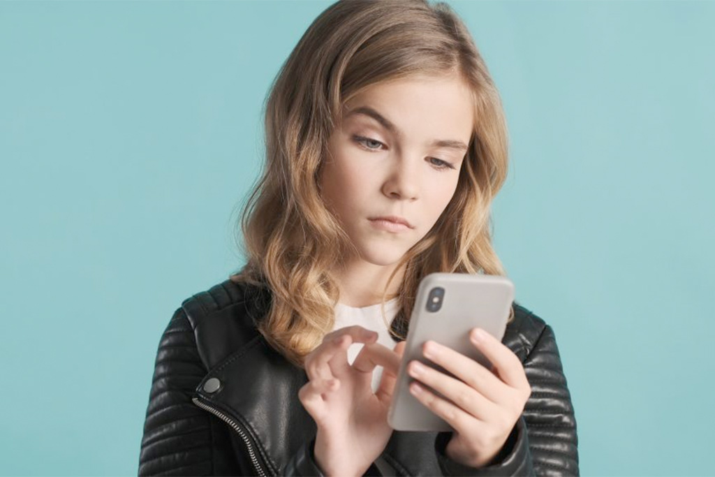 Langhaariges Mädchen vor blauem Hintergrund tippt mit skeptischem Blick auf ihr Smartphone.