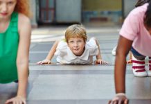 ein auf dem Bauch liegender Junge vor einem Liegestütz in einer Sporthalle lächelt mit erhobenem Kopf in die Kamera.