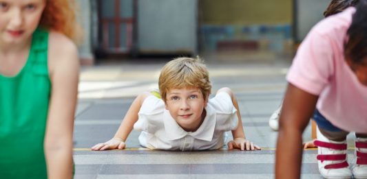 ein auf dem Bauch liegender Junge vor einem Liegestütz in einer Sporthalle lächelt mit erhobenem Kopf in die Kamera.