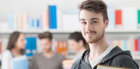 Ein Student schaut in die Kamera, hinter ihmverschwommen eien Lerngruppe in einer Bibliothek