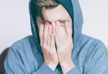 Junger Mann in hellblauem Hoodie sitzt auf einem Sofa und drückt die Finger ins Gesicht.