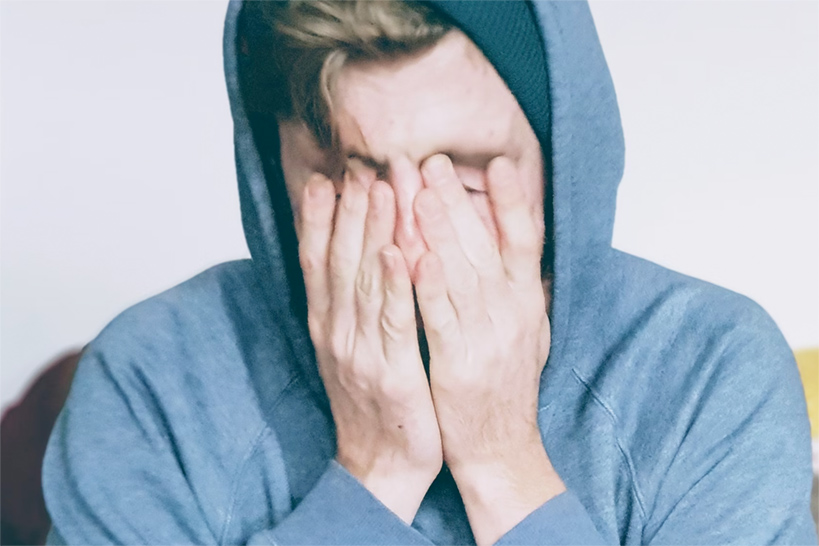Junger Mann in hellblauem Hoodie sitzt auf einem Sofa und drückt die Finger ins Gesicht.