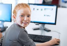 Jnnges Mädchen mit roten Haaren und blauweiß gestreiftem Pulllover blickt über die Schulter vom Bildschirm auf.