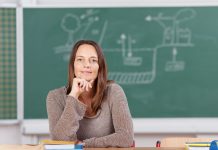 EIne Frau in graubraunem Pullover, mit langen, roten Haaren, das Kinn an die stifthaltende Hand gestützt sitzt an einem Pult. Hinter ihr verschwommen eine Tafel mit einer technische anmutenden Zeichnung.